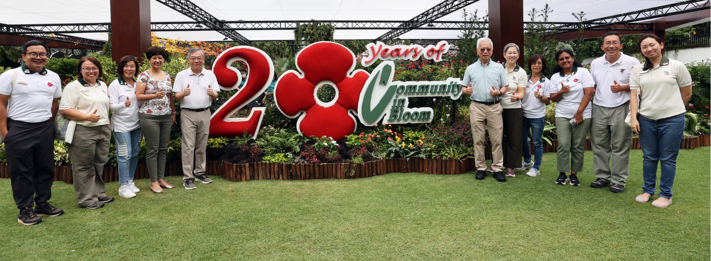 A group of people stands before a "20 Years of Community in Bloom" sign in a garden.